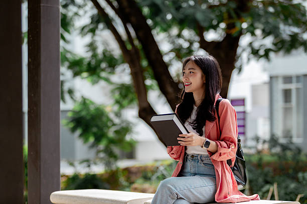 istock Young Asian woman is holding a book and smiling while sitting on a bench on a university campus 2209182255