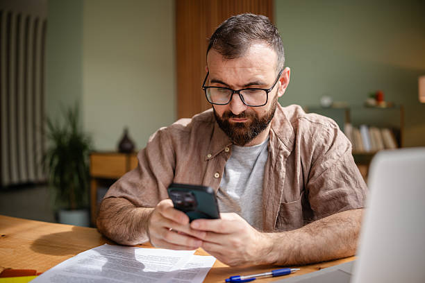 istock Working from home, a young man engages with his phone while surrounded by papers and a laptop in a cozy apartment setting 2209185965