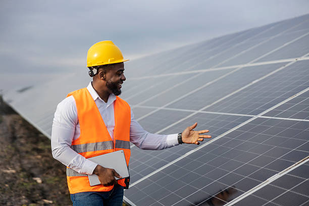 istock Young happy black engineer in protective work wear standing at solar farm with tablet in hands and showing at solar panels. 2209186891