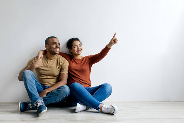 istock Young black couple embracing and pointing up while sitting on floor 2209194863