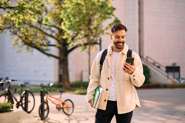 istock Smiling student walking in university campus using smartphone and carrying books 2209196786