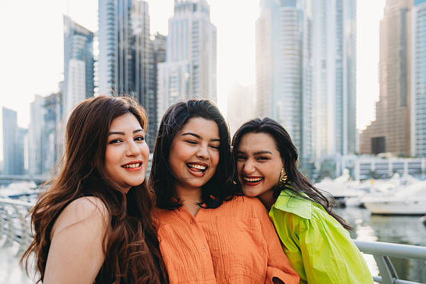 Three female friends are enjoying their vacation in Dubai Marina, UAE, laughing and posing for a picture with the cityscape in the background