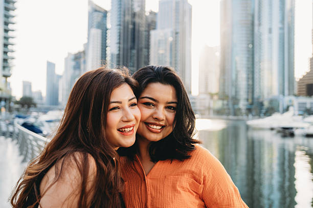 Two young women smiling and embracing each other, enjoying their time together in the vibrant Dubai Marina, surrounded by stunning skyscrapers