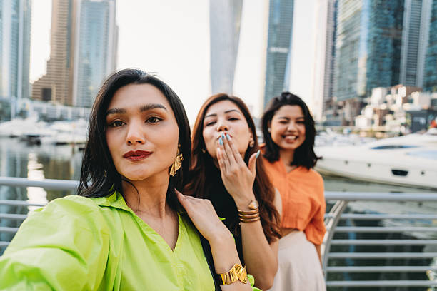 Three young female tourists are taking a selfie in Dubai Marina, with skyscrapers and yachts in the background