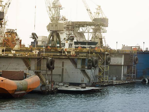 An offshore oil platform docked at a shipyard with cranes, support vessels, containers, and workers. The structure features yellow railings, massive tires, and maintenance crew on deck.