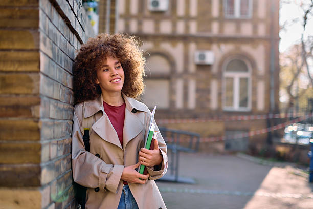 istock Smiling student leaning against brick wall holding books and looking away 2209213140