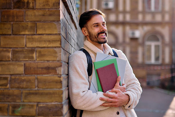 istock Smiling student leaning against brick wall, holding books and looking away 2209213182