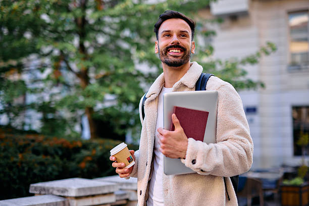 istock Smiling student walking outdoors holding laptop, book and takeaway coffee 2209213281