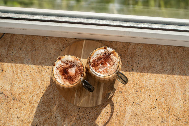 two frothy cappuccino mugs with cocoa dusting on a wooden tray by a sunlit window, casting soft shadows on a marbled surface, evoking a cozy morning ambiance. windowsill, inviting, aroma