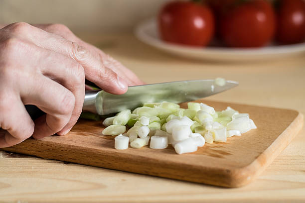 Men's hands are cutting green onions on a kitchen board. Tomatoes in the background create a bright culinary atmosphere with an emphasis on onions. Culinary still life.