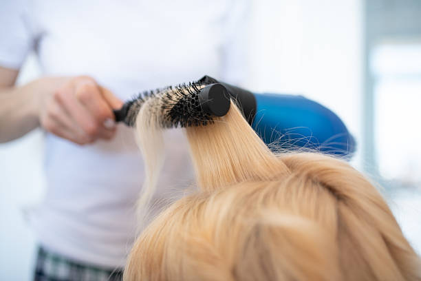 Close-up of a blonde woman’s head as a hairdresser styles her freshly washed hair with a round brush and blow-dryer.