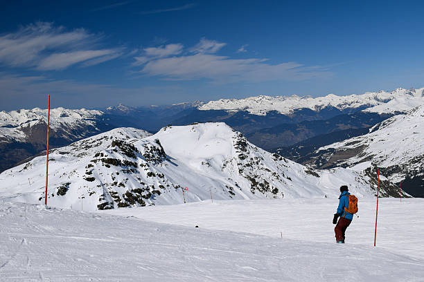 Les Menuires, France - March 14 2024: Man snowboarding down a ski slope overlooking snowcapped mountain peaks of the Three Valleys, France. View of snowy mountain valley from Les Menuires ski resort.