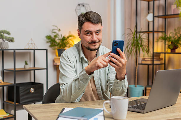istock Happy young man freelancer sitting at table using smartphone texting smiling at home apartment 2209259303