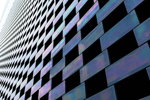 This image showcases a segment of a modern building's facade, characterized by a mesmerizing pattern of alternating black panels and iridescent tiles. The iridescent tiles shimmer with a spectrum of colors, reflecting the sky and surroundings in a dance of light and shadow. The stark contrast between the dark panels and the vibrant hues of the iridescent tiles creates a visually striking rhythm across the building's surface. The perspective of the photo, taken from a low angle, enhances the building's towering presence and the geometric precision of its design. This architectural feature not only serves an aesthetic function but also demonstrates the innovative use of materials and the dynamic interaction between structure and environment. The image captures the essence of contemporary design, where form and function are seamlessly integrated to create visually compelling and interactive spaces.