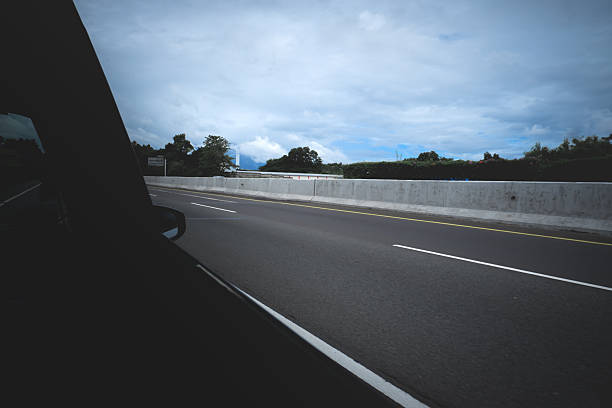 Scenic view of an empty highway captured from a moving car window, showing roadside trees, concrete barrier, and a partly cloudy blue sky. Peaceful road trip atmosphere.