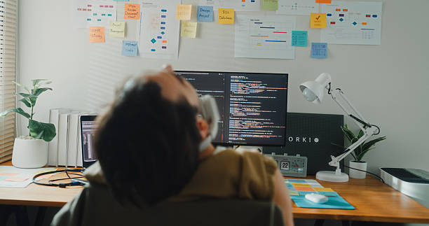 istock Young gay programmer stretches to ease backache while working on AI prompts and web designs coding user experience on laptop in bedroom at home. Software developer, artificial intelligence. 2209310215