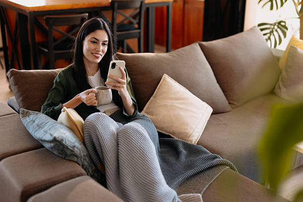 istock Young woman relaxing on the sofa, using smartphone and drinking tea 2209311858