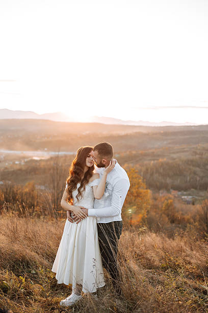 A couple is standing on a hillside, with the sun setting behind them