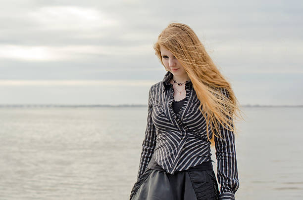 Young female with long blonde hair, wearing striped blouse, walks by calm seaside. Overcast sky gives muted tones to serene setting. Gentle breeze adds dynamic movement to hair