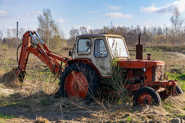 istock An old, rusty tractor and a backhoe loader stand in an abandoned field. The vehicle is overgrown with vegetation, indicating long-term disuse. In the background is a spring landscape with trees without leaves. 2209325270