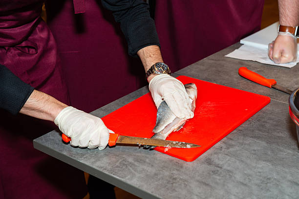Person in gloves using a knife to fillet a raw fish on a red cutting board during a cooking class or kitchen prep.