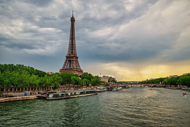 Eiffel Tower by the Seine River in Paris at sunset with dramatic clouds and vibrant greenery.