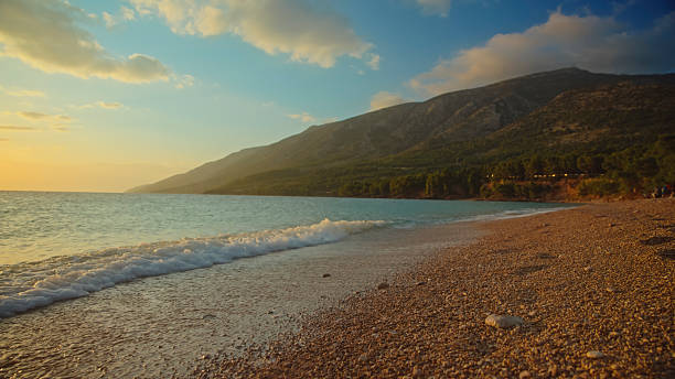 Panoramic view of crystal-clear waves washing onto pebbled shoreline at Zlatni Rat Beach, Croatia. Mountainous coastline stretches beneath golden sunset sky with scattered clouds.