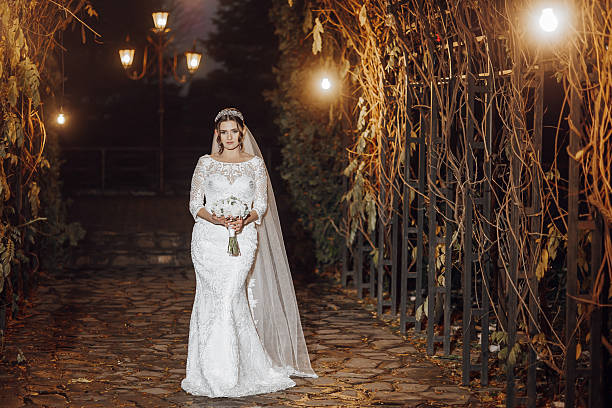 A bride is walking down a stone path with a bouquet of flowers in her hand. The scene is set in a dimly lit area, with a few lights illuminating the path. The bride is wearing a white dress and a veil