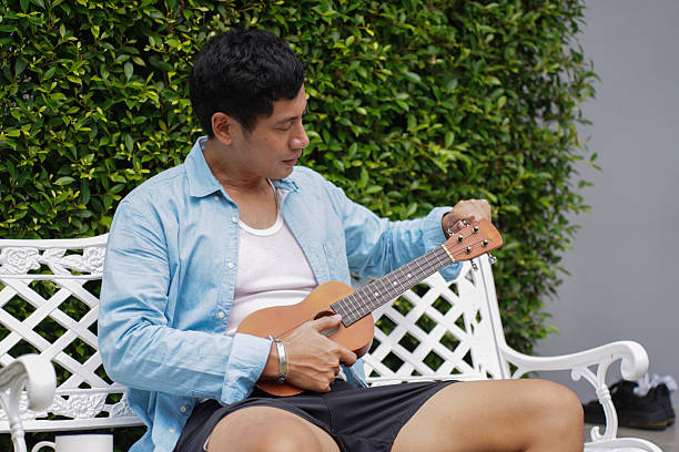 young asian man sitting outdoors on white bench tuning ukulele, enjoying peaceful and relaxing morning activity