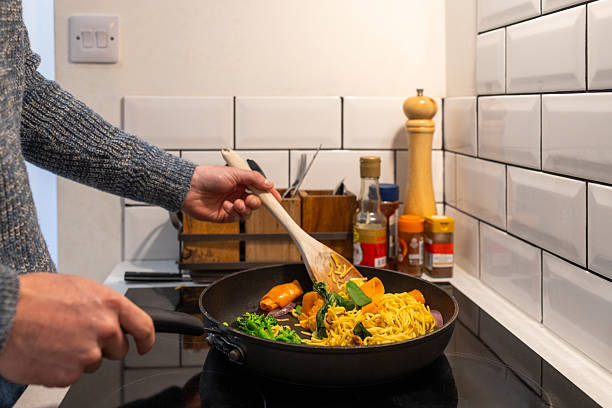A close-up of an unrecognisable man cooking a fresh stir-fry noodle dish in a cozy kitchen. Colorful vegetables and noodles are being stirred in a frying pan on the stove, with spices and sauces in the background. \n\nVideos of this scenario are also available