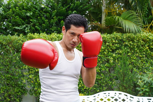 Asian male boxer in red gloves posing strong and determined in garden, wearing white tank top, showing fighting stance and confidence