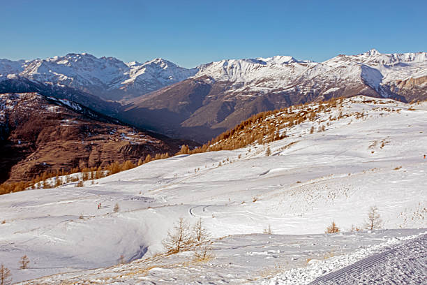 A wide-angle landscape shot of a massive, snow-blanketed mountain range beneath a brilliant, deep blue sky. The foreground highlights the unique wind-blown textures and ripples in the snow, leading the viewer's eye toward several sharp, sunlit peaks. The image evokes a sense of cold, quiet majesty and the vastness of the alpine wilderness, perfect for travel, adventure, and nature-themed projects.