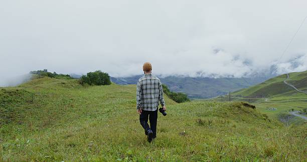 Russia, Caucasus, Elbrus. A guy in a hat with a camera in his hand walks along a grassy mountain slope. Clouds hide the tops of the mountains. Daylight video
