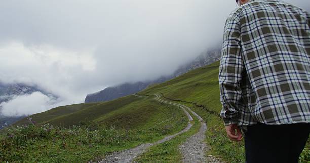 Russia, Caucasus, Elbrus. A young fashionably dressed man in a hat walks along a mountain path along a mountain slope overgrown with grass in daylight. The tops of mountains are covered with clouds