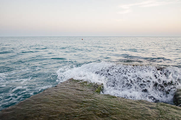Waves crash against moss-covered rock as sun sets over calm ocean. Light reflects softly on water, creating peaceful atmosphere. Horizon blends into pastel sky