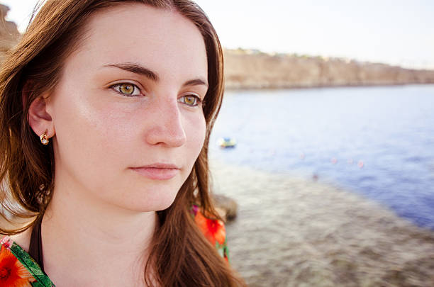 Young woman with long brown hair gazes thoughtfully at sea. Sunshine reflects on clear water cliffs in distant background add depth. Calm, contemplative mood