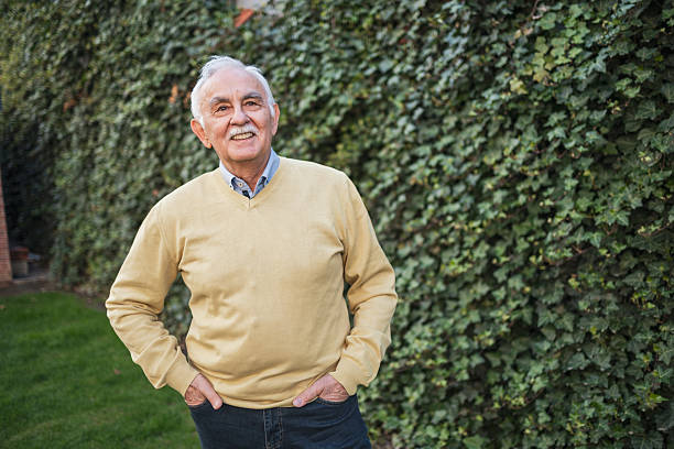 A cheerful elderly man with a mustache smiles outdoors beside a lush green hedge. His demeanor exudes happiness and positivity, conveying the essence of joy and connection with nature.