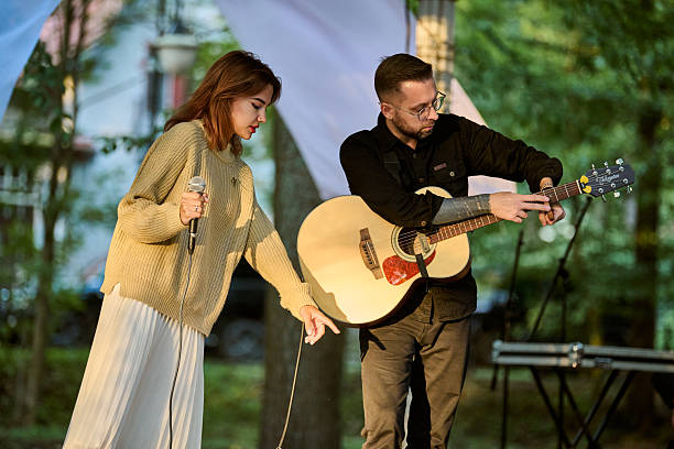 Kaliningrad, Russia - 10.08.2024 - Young woman sings beside male guitarist in sunlit park. Trees and soft light create serene atmosphere
