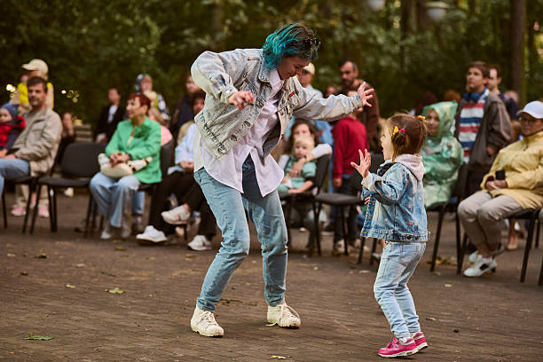 Kaliningrad, Russia - 11.08.2024 - Adult female with blue hair and child, both caucasian, joyfully dancing in park. Casual crowd, natural light, green backdrop