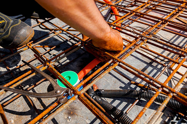 Worker's hands as he assembly protective ribbed plastic hoses, for cables, placing and connect them to distribution box, under rusty rebar, prepared for pouring concrete over them.