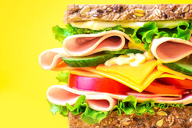 Big layered sandwich with ham, lettuce, cheddar cheese, tomato and cucumber on bright yellow background, extremely close up, copy space. Healthy lunch sandwich with wholegrain bread, bacon, tomato, cheese and sauce.