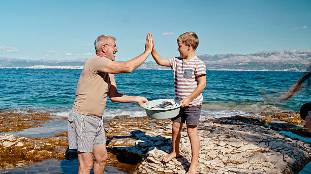 Caucasian grandfather and grandson bond while gutting fish on rocky seaside terrain under clear skies. The boy holds a bowl of fresh fish, sharing a cheerful high-five with his grandfather in the bright sunlight.