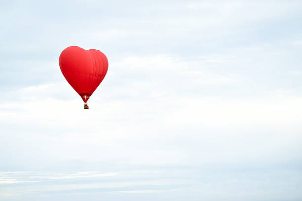 This image captures a striking red hot air balloon uniquely designed in the shape of a heart, floating gracefully in a cloudy sky. The background features soft hues of white and blue, adding depth and serenity to the composition. A small basket is attached to the base of the balloon, suggesting its capability for carrying passengers. This visually appealing and unusual design adds a romantic and adventurous charm, making it ideal for travel promotions, themed events, or advertising creative experiences.