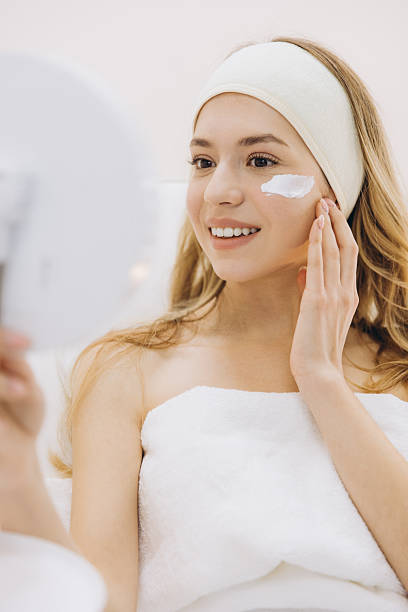 istock Smiling woman applying face cream while looking in a mirror, wrapped in a towel, showcasing a skincare routine in a spa like environment 2209401830