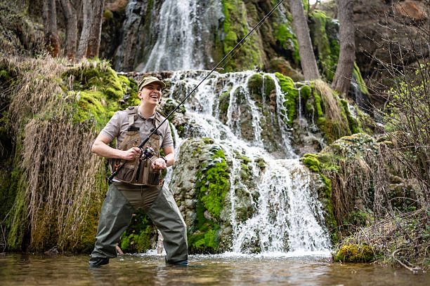 An adventurous man is fly fishing near a majestic waterfall surrounded by lush greenery and flowing water. The serene natural environment captures outdoor exploration, peace, and enjoyment of nature.
