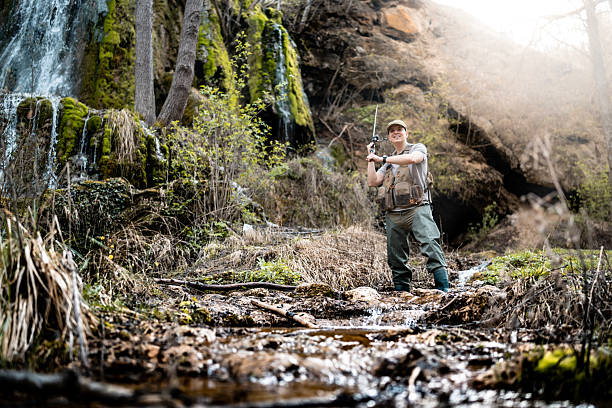 An adventurous fly fisherman standing in a lush forest creek, holding a rod and smiling brightly. The scene features a tranquil flowing stream and greenery creating a peaceful and captivating environment.