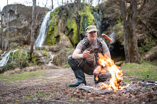 A man wearing outdoor gear cooks a sausage over a campfire beside a waterfall surrounded by lush greenery, highlighting outdoor adventure and connection with nature.