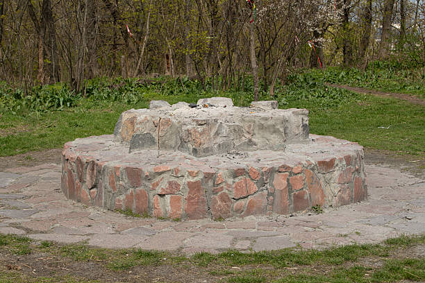 A stone fire pit sits prominently in a park, surrounded by fresh greenery and trees. The area is tranquil, ideal for gatherings and enjoying nature in spring.