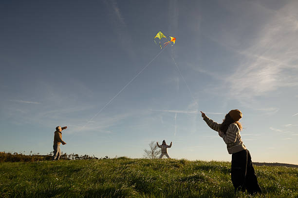 Father and two daughters enjoying a sunny day in a vibrant green field, flying colorful kites and pointing excitedly at the fluffy clouds in the bright blue sky