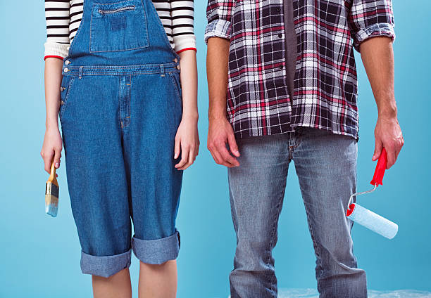 Young adult couple standing against blue wall and holding painting materials - paintbrush and paint roller - in their hands.